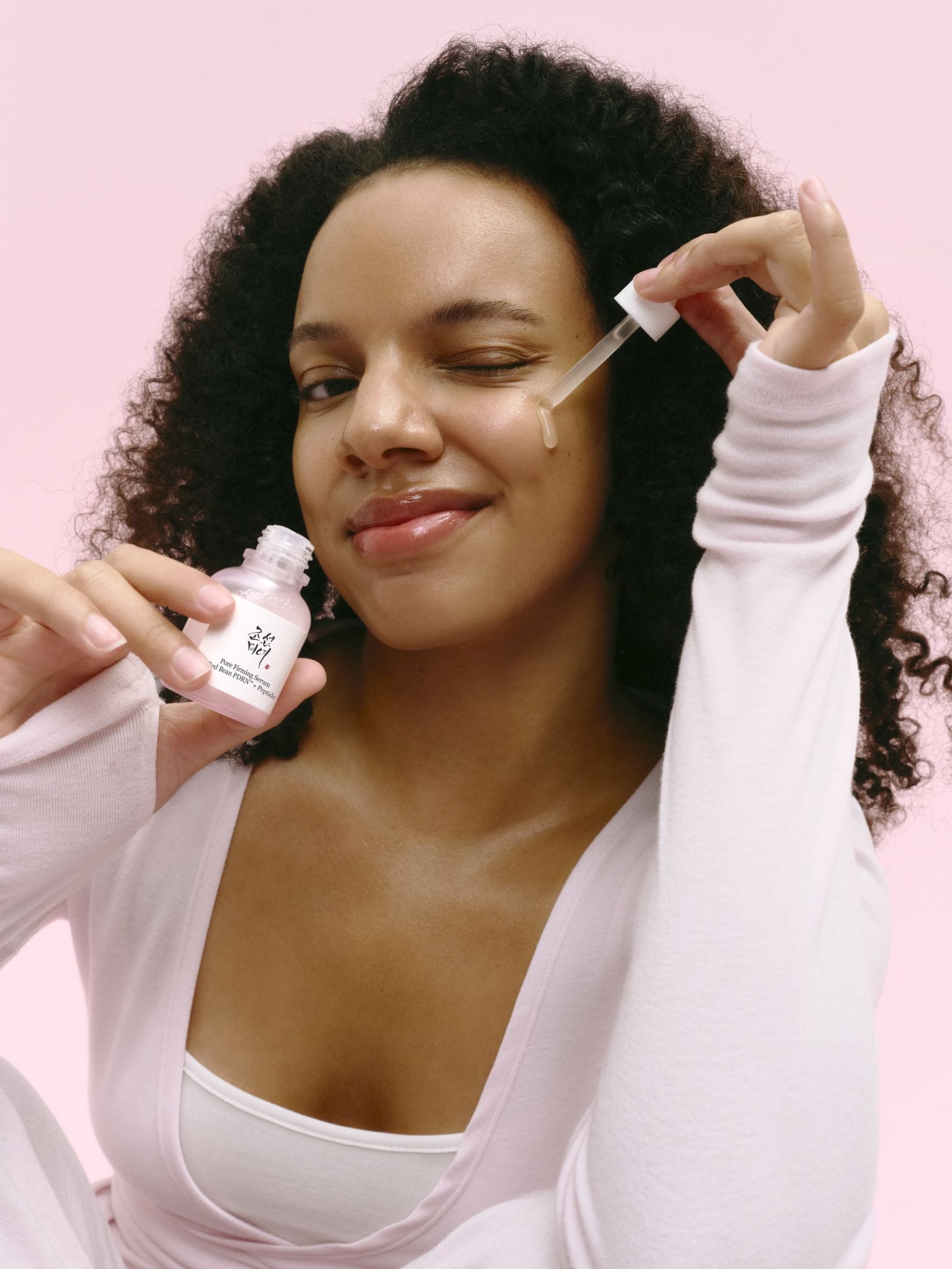A woman with curly black hair applies a skincare serum from a small pink bottle using a dropper, set against a solid pink background.