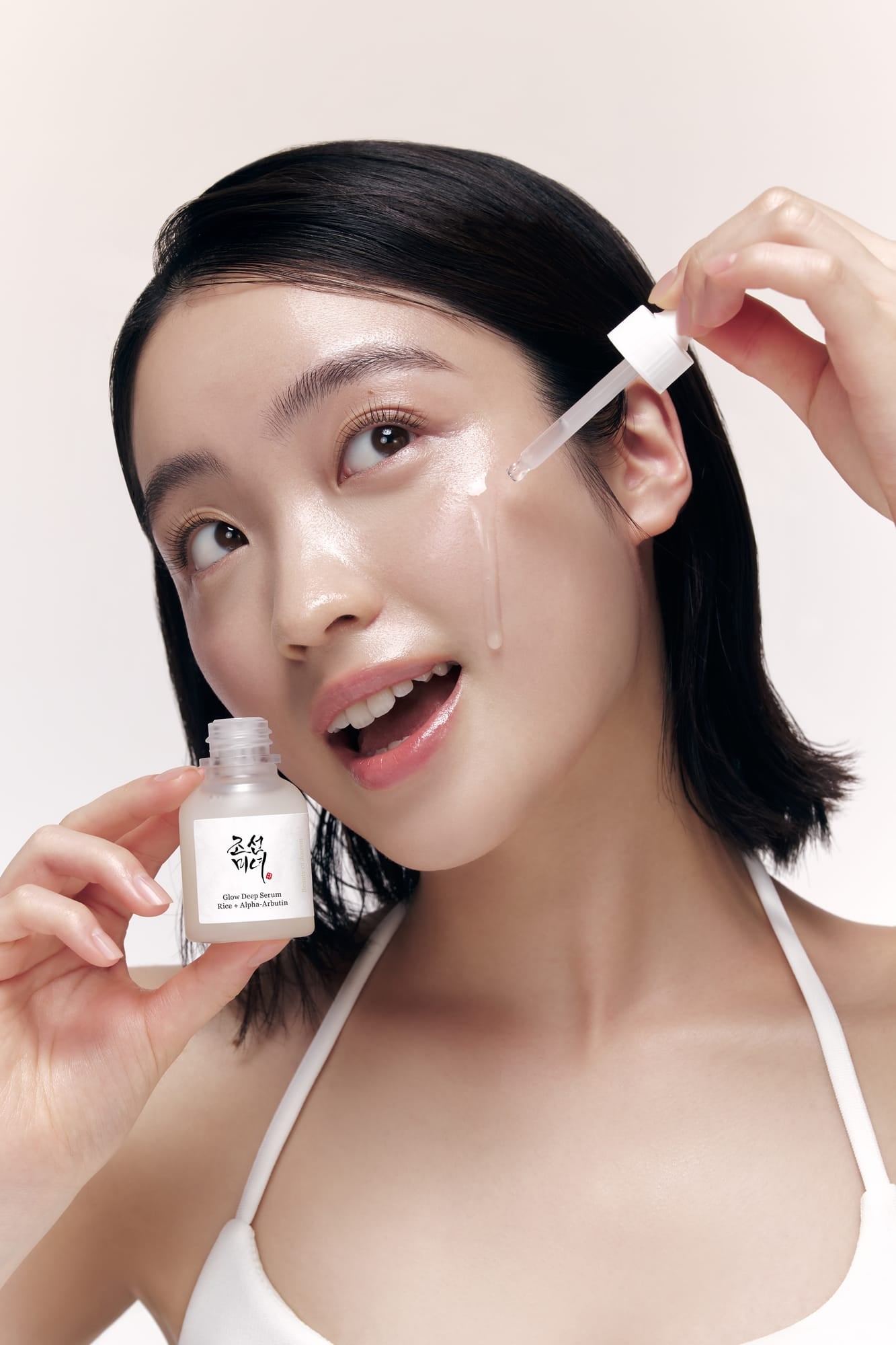 A woman with short dark hair applies a Glow Deep Serum from a dropper to her cheek while holding a small white bottle with a Korean label. She is wearing a white spaghetti-strap top against a soft light beige studio background.